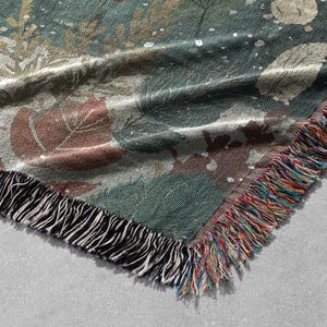 May include: A woven throw blanket with a botanical design. The blanket features a teal background with brown and white leaf and floral patterns. The edge is finished with a fringe of black and multicolored threads.