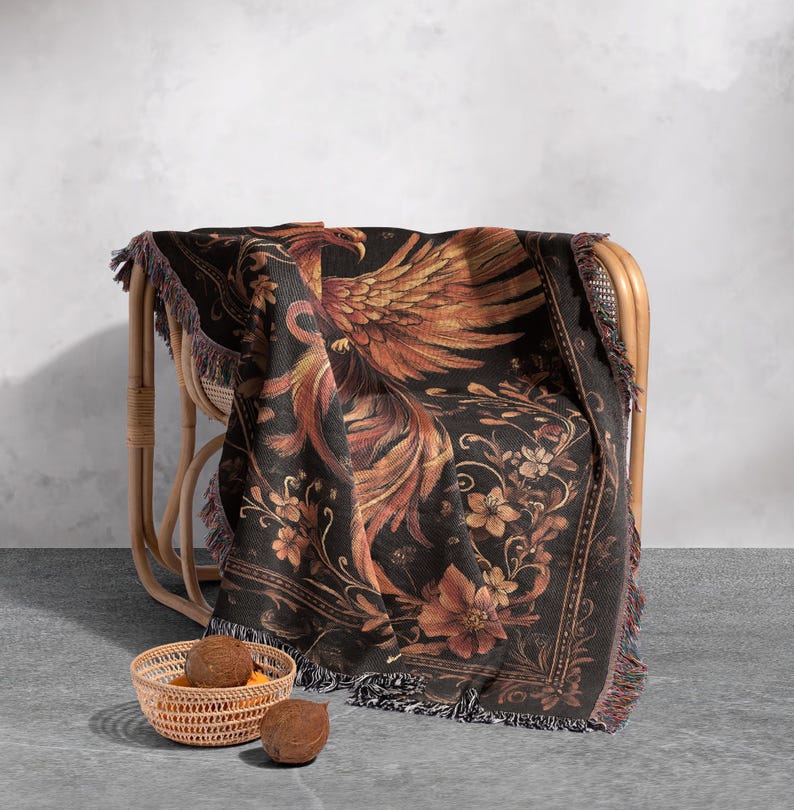 May include: A black woven throw blanket with a gold and orange phoenix design and floral accents. The blanket has fringe on all sides and is draped over a wicker chair.