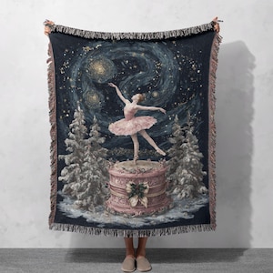 May include: A woven throw blanket featuring a ballerina in a pink tutu, dancing on a decorative pink platform. The background depicts a starry night sky and snow-covered trees. The blanket has a dark blue border with fringe.