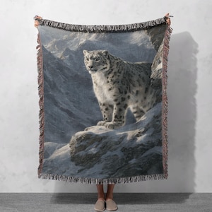 May include: A woven throw blanket featuring a snow leopard in a mountainous, snowy landscape. The blanket has a fringed edge and a detailed illustration of the animal. The color palette includes shades of gray, white, and brown.