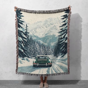 May include: A woven throw blanket featuring a vintage green car driving through a snowy mountain landscape. The car is driving on a winding road with snow-covered trees on either side. The mountains in the background are covered in snow.
