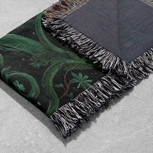 May include: A folded black throw blanket with a green botanical design. The design features swirling vines and leaves. The blanket has a fringed edge with black, white, and multicolored threads. The reverse side is a solid gray.