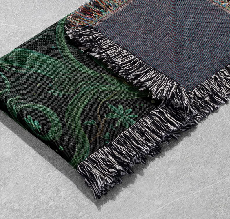 May include: A folded black throw blanket with a green botanical design. The design features swirling vines and leaves. The blanket has a fringed edge with black, white, and multicolored threads. The reverse side is a solid gray.