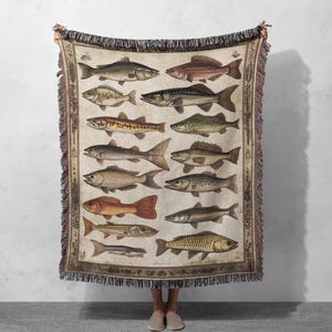 Puede incluir: Una manta de tejido de tapicería con una ilustración vintage de 14 tipos diferentes de peces. Los peces están representados en varios tonos de marrón, verde y naranja, con detalles intrincados y texturas realistas. El fondo es una tela de color crema con un patrón sutil.