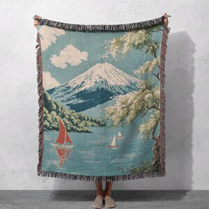 Könnte beinhalten: Eine gewebte Decke mit einer Berglandschaft mit Segelbooten auf einem See. Die Decke hat einen blauen Hintergrund mit einem Fransenrand. Die Illustration zeigt einen schneebedeckten Berg, Bäume und Wolken.