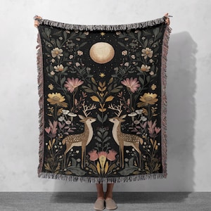 May include: Black woven throw blanket with a floral and deer design. The blanket features two deer facing each other, a full moon, and floral elements. The blanket has a fringed edge.