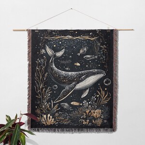 May include: A woven wall tapestry featuring a black and white illustration of a whale swimming in a starry ocean. The whale is surrounded by seaweed, coral, and small fish. The tapestry has a fringed edge.