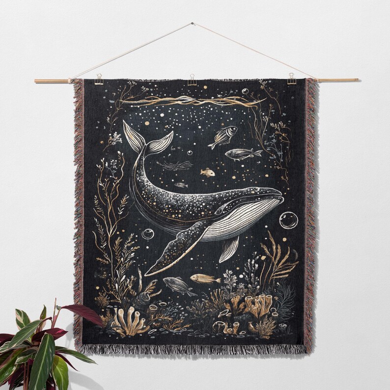May include: A woven wall tapestry featuring a black and white illustration of a whale swimming in a starry ocean. The whale is surrounded by seaweed, coral, and small fish. The tapestry has a fringed edge.
