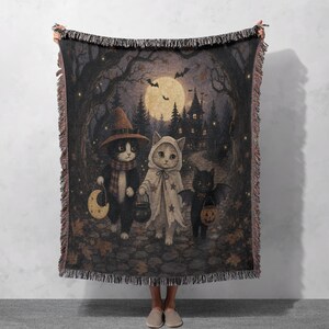 Könnte beinhalten: Eine gewebte Wohndecke mit einer Halloween-Szene, die drei Katzen in Kostümen zeigt. Eine Katze trägt einen Hexenhut, eine ein Geisterkostüm und eine Fledermausflügel. Sie laufen auf einem Pfad auf ein dunkles Haus unter einem Vollmond zu.
