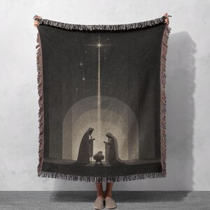 Könnte beinhalten: Eine dunkelgraue gewebte Wohndecke mit einer Darstellung der Geburt Christi. Das Design zeigt Silhouetten von Maria, Josef und dem Jesuskind unter einem hellen Stern. Die Decke hat einen Fransenrand.