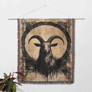 Pu&ograve; includere: Un arazzo da parete tessuto con un'illustrazione in bianco e nero di una testa di capra con corna all'interno di un cerchio. Lo sfondo &egrave; di colore beige con motivi geometrici in bianco e nero.