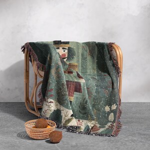 May include: A woven throw blanket draped over a wicker chair. The blanket features a detailed illustration of a nutcracker soldier in shades of green, red, and beige. The edges are finished with a fringed trim.