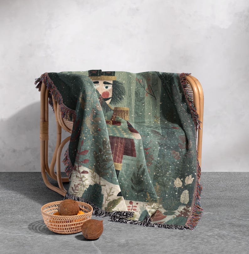 May include: A woven throw blanket draped over a wicker chair. The blanket features a detailed illustration of a nutcracker soldier in shades of green, red, and beige. The edges are finished with a fringed trim.