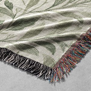 Op de afbeelding: Een geweven sprei met een botanisch bladerpatroon in groen- en cr&egrave;metinten. De sprei heeft een gefranjerde rand met een mix van kleurrijke draden. De hoek van de sprei is zichtbaar en toont de textuur en het ontwerp.
