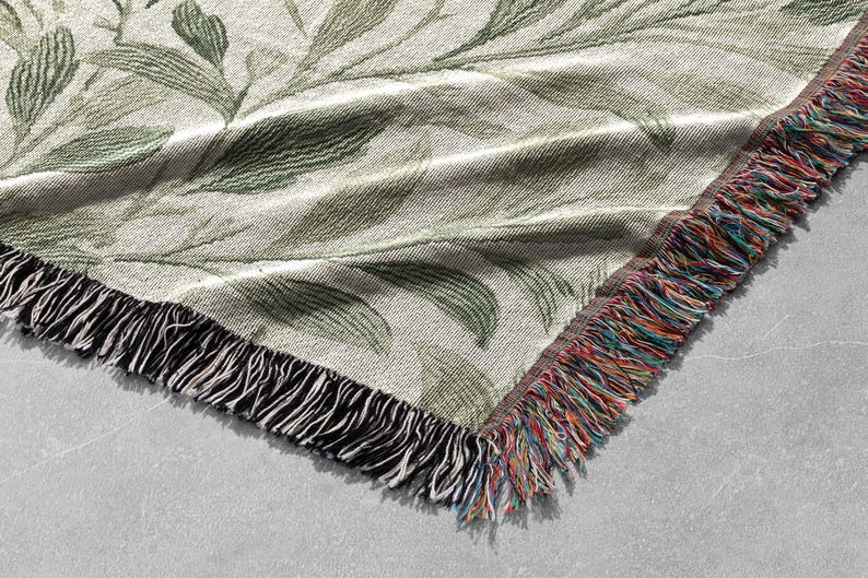 Op de afbeelding: Een geweven sprei met een botanisch bladerpatroon in groen- en cr&egrave;metinten. De sprei heeft een gefranjerde rand met een mix van kleurrijke draden. De hoek van de sprei is zichtbaar en toont de textuur en het ontwerp.