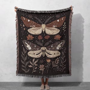 May include: A woven tapestry throw blanket with a dark brown background featuring two moths with brown, white, and black wings. The moths are surrounded by small, stylized flowers and leaves in shades of brown, pink, and white.