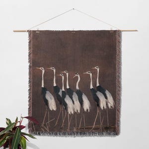 Pode incluir: Uma tape&ccedil;aria de parede tecida com uma linha de sete gruas cinzas e brancas com pesco&ccedil;os longos e bicos pretos, em p&eacute; em fila sobre um fundo castanho.