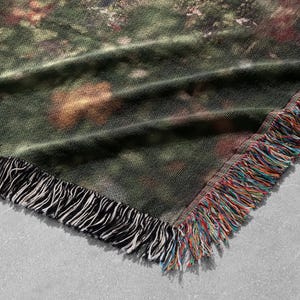 Pu&ograve; includere: Una coperta tessuta con un disegno di scena naturale. La coperta presenta una tavolozza di colori verde, marrone e rosso con un bordo a frange nero e una frangia multicolore. La coperta &egrave; piegata, mostrando l'angolo.