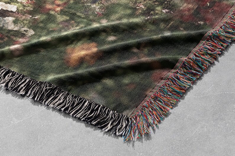 Pu&ograve; includere: Una coperta tessuta con un disegno di scena naturale. La coperta presenta una tavolozza di colori verde, marrone e rosso con un bordo a frange nero e una frangia multicolore. La coperta &egrave; piegata, mostrando l'angolo.