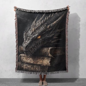 May include: A woven throw blanket featuring a dark grey dragon's head with a glowing yellow eye, resting on a stack of old books. The dragon's head is the main focus of the image, with the books providing a backdrop.