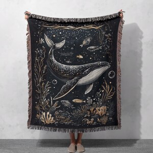 May include: A woven tapestry throw blanket featuring a black and white illustration of a whale swimming in a starry ocean with gold accents. The whale is surrounded by coral, seaweed, and fish.