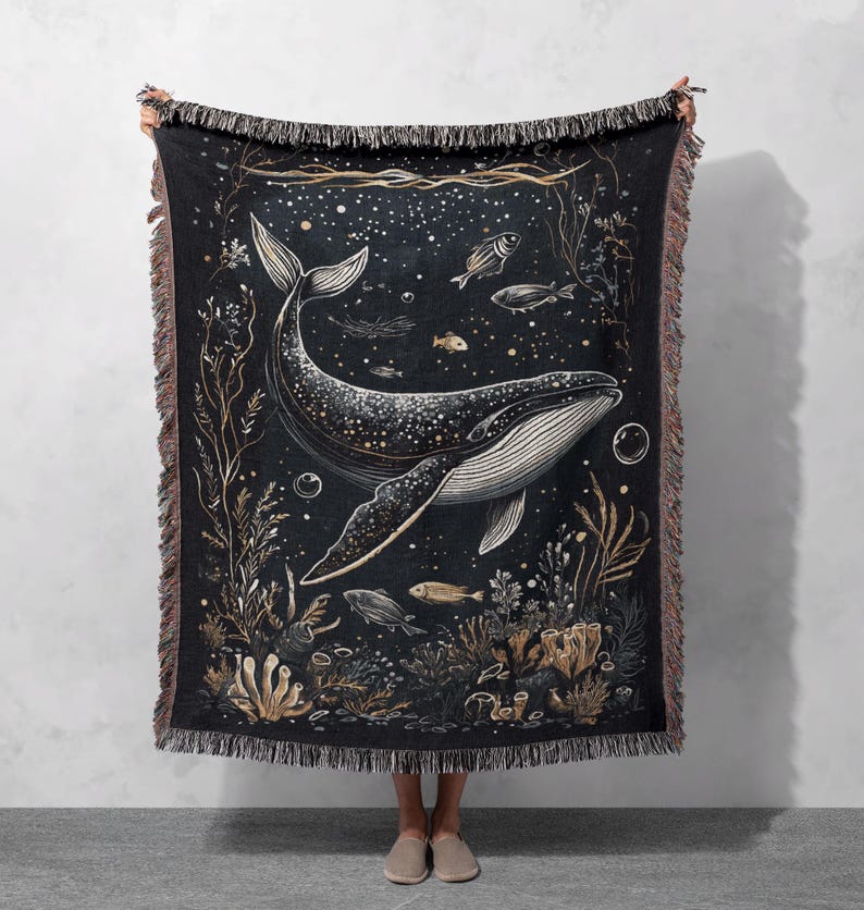 May include: A woven tapestry throw blanket featuring a black and white illustration of a whale swimming in a starry ocean with gold accents. The whale is surrounded by coral, seaweed, and fish.