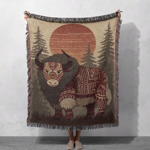 May include: Woven throw blanket featuring a stylized yak design. The blanket showcases a yak with intricate patterns in shades of brown, red, and beige. The background depicts a sunset scene with trees. The blanket has a fringed edge.