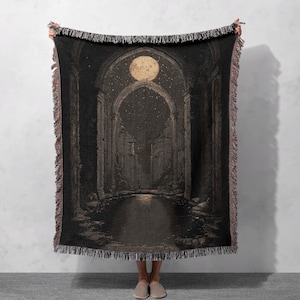 May include: A woven throw blanket featuring a nighttime scene with a full moon, stars, and an arched architectural structure. The blanket has a dark color palette with a fringed edge. The design evokes a sense of mystery and fantasy.