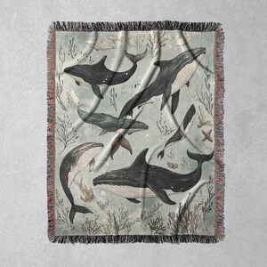 May include: A woven tapestry featuring a blue and white design of whales swimming in a sea of coral and seaweed. The tapestry has a fringed border.