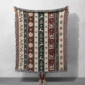 Pode incluir: Uma manta tecida com um padrão de riscas verticais e motivos festivos, incluindo poinsétias, veados a saltar, pinheiros e pequenas cabanas. O design incorpora tons de vermelho escuro, verde floresta e creme com extremidades franjadas.