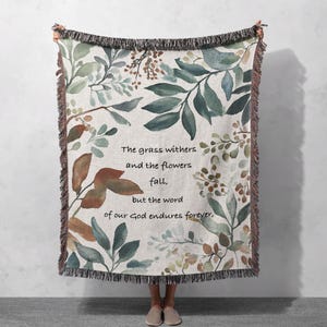 May include: A woven throw blanket with a beige background and a watercolor floral pattern in shades of green, brown, and white. The blanket features a quote: "The grass withers and the flowers fall, but the word of our God endures forever."