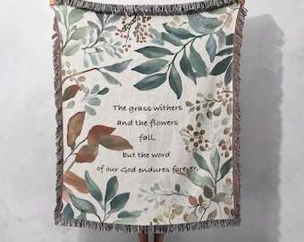 May include: A woven throw blanket with a beige background and a watercolour floral pattern in shades of green, brown, and white. The blanket features a quote: "The grass withers and the flowers fall, but the word of our God endures forever."