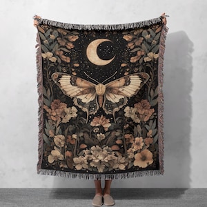 May include: A woven tapestry throw blanket with a dark background featuring a large, detailed butterfly with brown and white wings. The butterfly is centred in a field of flowers with a crescent moon and stars in the background.