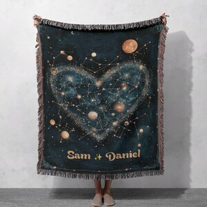 May include: A woven throw blanket with a dark blue background featuring a constellation design in the shape of a heart. The heart is filled with stars and planets. The names "Sam + Daniel" are written in gold lettering at the bottom of the blanket.