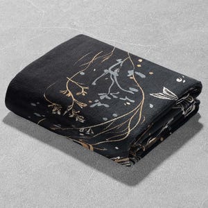 May include: A black fabric throw blanket with a gold floral pattern. The blanket is folded in thirds and is lying on a gray surface.