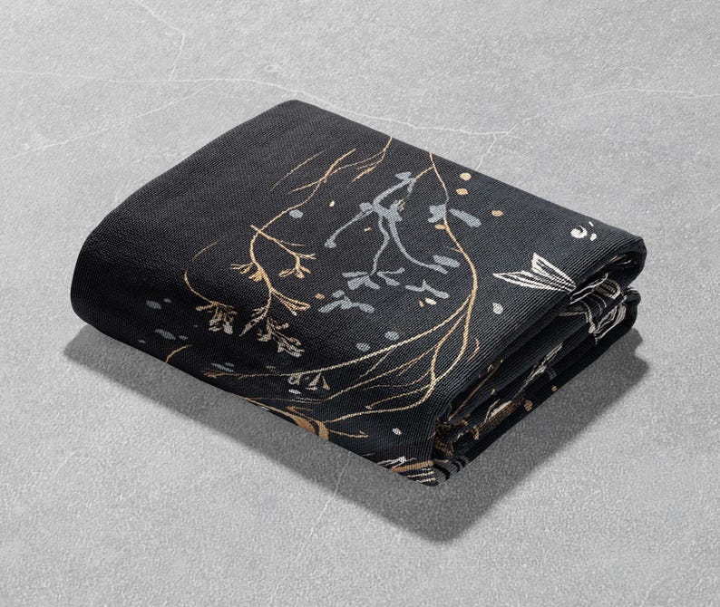 May include: A black fabric throw blanket with a gold floral pattern. The blanket is folded in thirds and is lying on a gray surface.