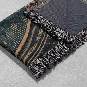May include: A folded woven blanket with a dark teal and tan design. The blanket has a fringed edge with black and white threads. The design features abstract lines and shapes. The blanket is made of a soft material.