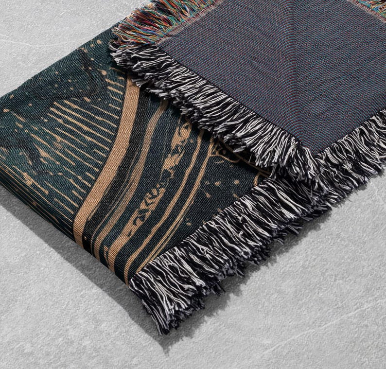 May include: A folded woven blanket with a dark teal and tan design. The blanket has a fringed edge with black and white threads. The design features abstract lines and shapes. The blanket is made of a soft material.