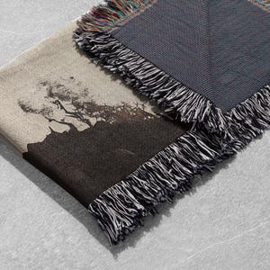May include: A woven throw blanket with a black and white image of a mountain range and trees. The blanket has a fringe border and is folded in half.