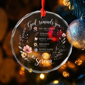 Peut inclure: Une décoration circulaire en acrylique transparent à facettes suspendue par un ruban rouge. Elle présente un motif de couronne florale entourant le texte God reminds you, You are chosen, Strong, Deeply loved, Precious, Never alone et le nom Serena.