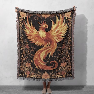 May include: A woven throw blanket featuring a detailed illustration of a phoenix with orange and yellow feathers against a black background. The phoenix is surrounded by floral designs in shades of orange, yellow, and brown.