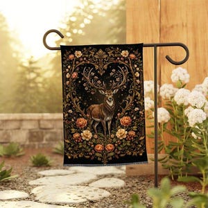May include: A rectangular garden flag featuring a central illustration of a deer with large antlers, surrounded by an ornate frame of blooming roses and foliage on a black background, suspended from a black metal garden flag stand.