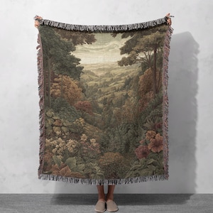 May include: A woven throw blanket featuring a detailed landscape scene with trees, hills, and flowers in shades of green, brown, and beige. The blanket has a fringed edge and is held up against a neutral background.