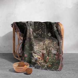 Pu&ograve; includere: Una coperta tessuta con una stampa di giardino panoramico, drappeggiata su una sedia di vimini. La coperta raffigura un sentiero di pietra attraverso un giardino con fiori rosa e fogliame verde. Ha un bordo a frange.