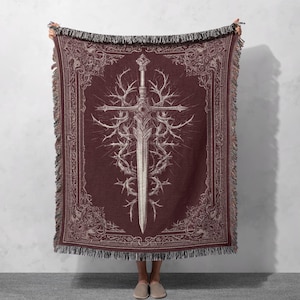 May include: A burgundy throw blanket with a detailed white illustration of a sword surrounded by thorny vines. The blanket has a decorative border and fringe along the edges, creating a gothic or fantasy theme.