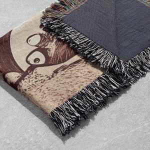May include: A woven tapestry throw blanket featuring a brown and white cartoon image of a creature with large eyes. The blanket has a black fringed border.