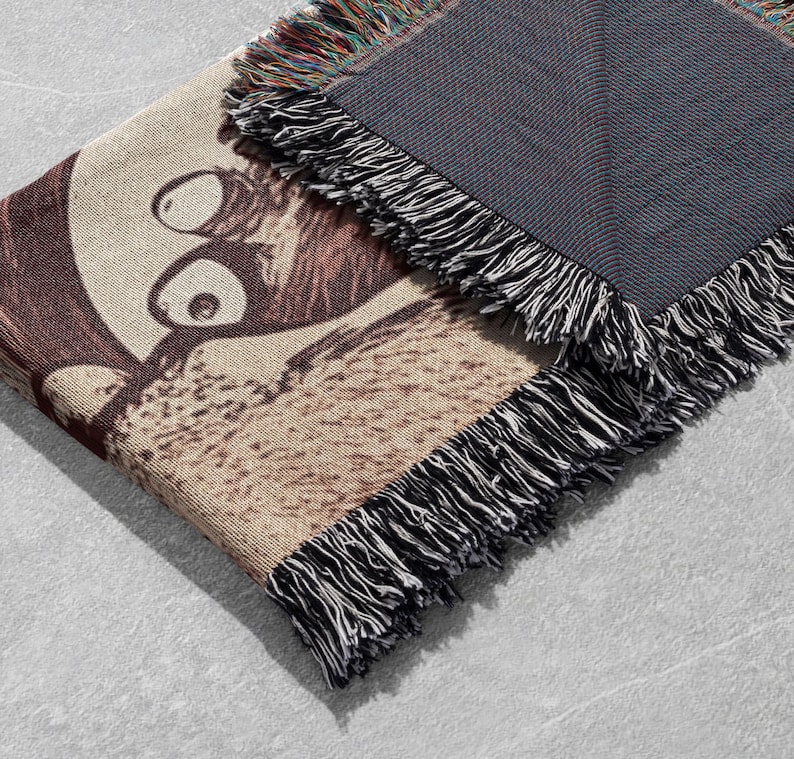 May include: A woven tapestry throw blanket featuring a brown and white cartoon image of a creature with large eyes. The blanket has a black fringed border.