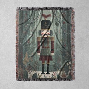 May include: A woven throw blanket featuring a whimsical illustration of a nutcracker soldier in a forest setting. The design includes a detailed nutcracker figure in a uniform, set against a backdrop of trees and falling snow. The blanket has a fringed edge.