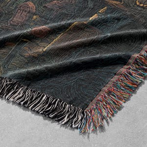 May include: A woven throw blanket with a dark background and a colorful, artistic design. The blanket has a fringed edge with a mix of colors, including red, orange, and blue. The design appears to be a painting.