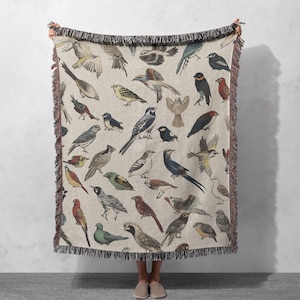 Puede incluir: Una manta tejida con un fondo beige y un patrón de pájaros coloridos. Los pájaros están representados en un estilo vintage y están dispuestos en un patrón repetitivo.